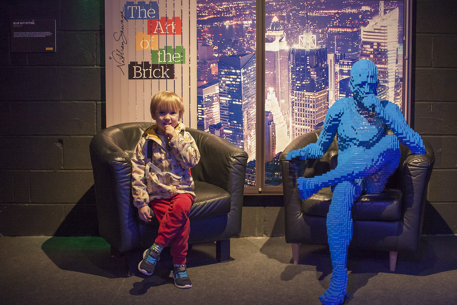 A boy copying the pose of a life size Lego man sitting in a chair