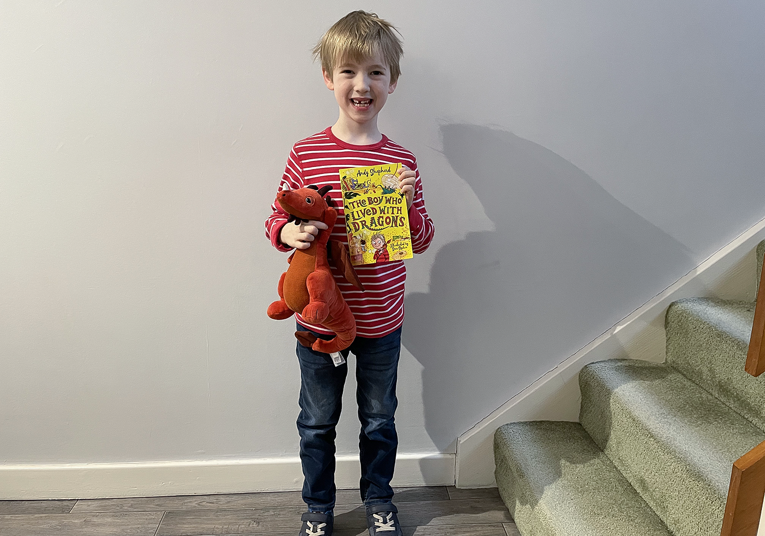 Gabe wearing a red stripy t-shirt and jeans, holding a red dragon stuffed toy and a copy of The Boy Who Lived with Dragons